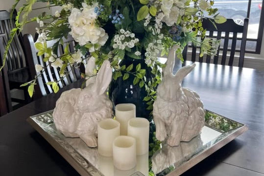 Decorative table setting with floral arrangement, rabbits, and candles on a mirror tray.