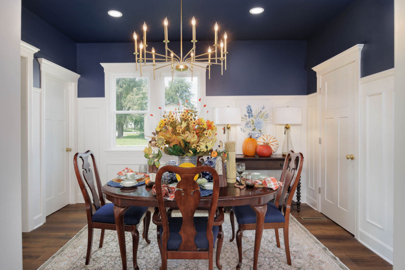 Dining room with a round table and chairs, decorated for fall with pumpkins and flowers, under a chandelier.