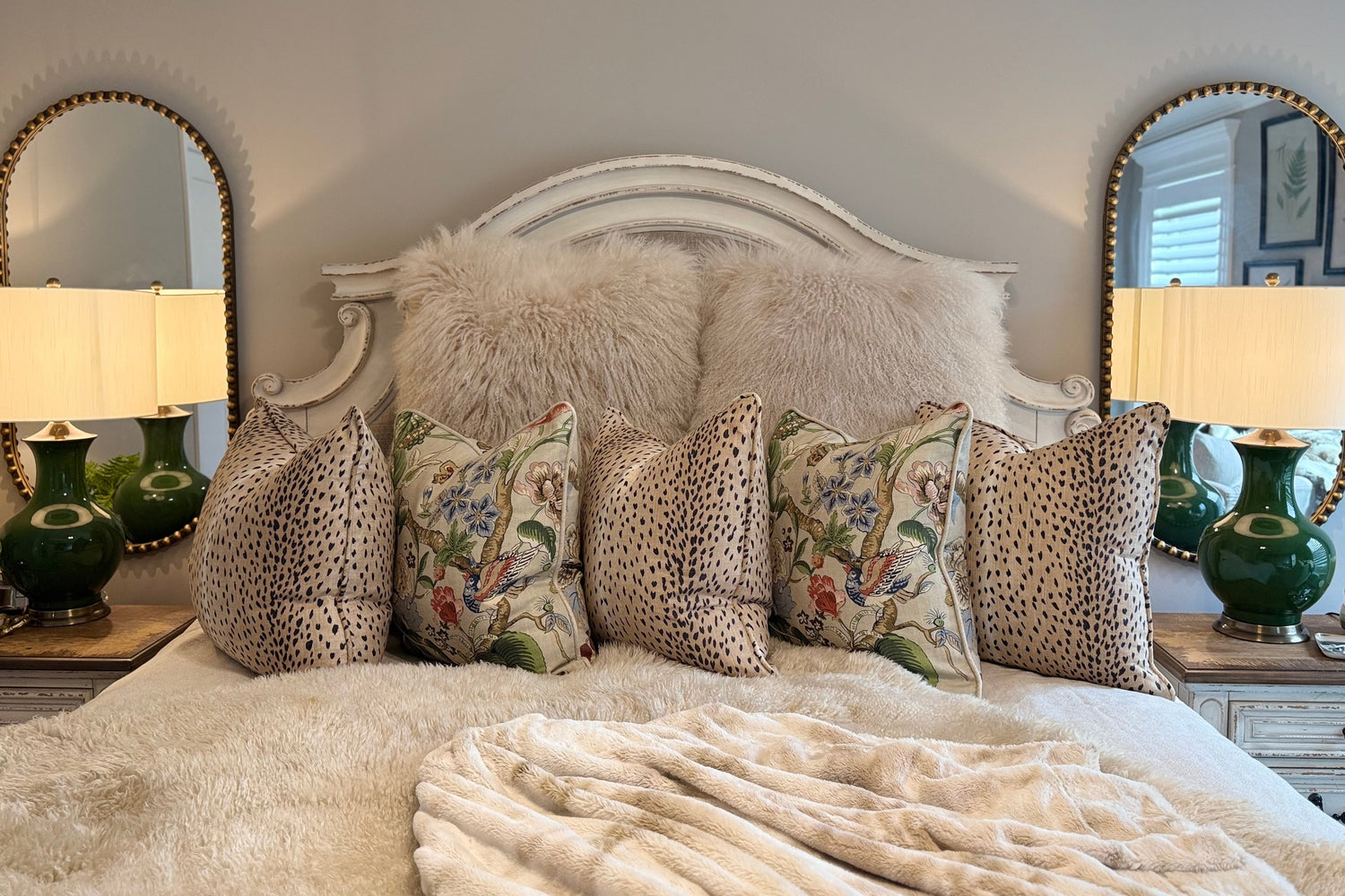 Beautifully layered bedding with cheetah pillows, floral printed pillows, faux fur, and classic linens. Nightstands with deep rich green lamps and matching golden studded mirrors.