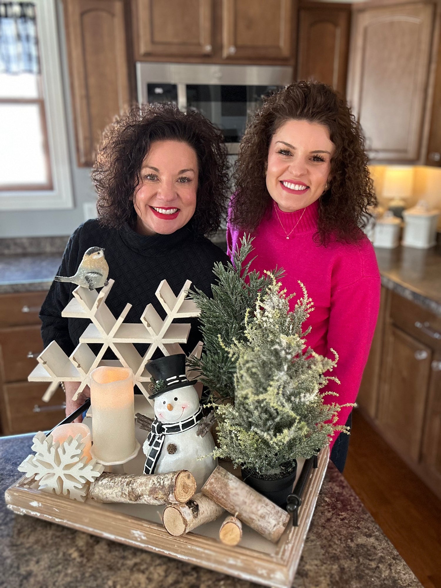 Two women standing in a kitchen with a decorative tray featuring a snowflake, candle, and small tree.