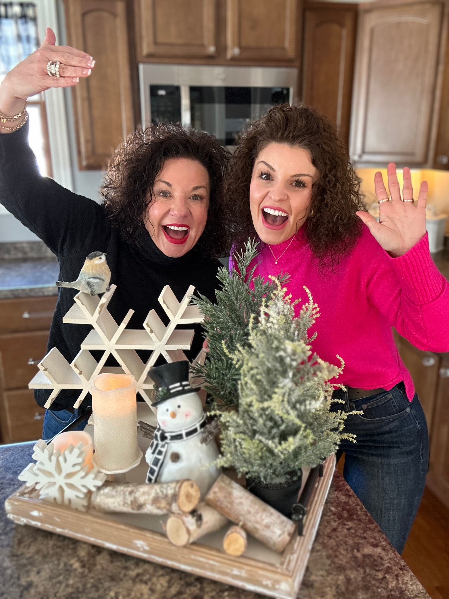 Two women in a kitchen with a decorative tray featuring a snowflake, candle, and small tree.