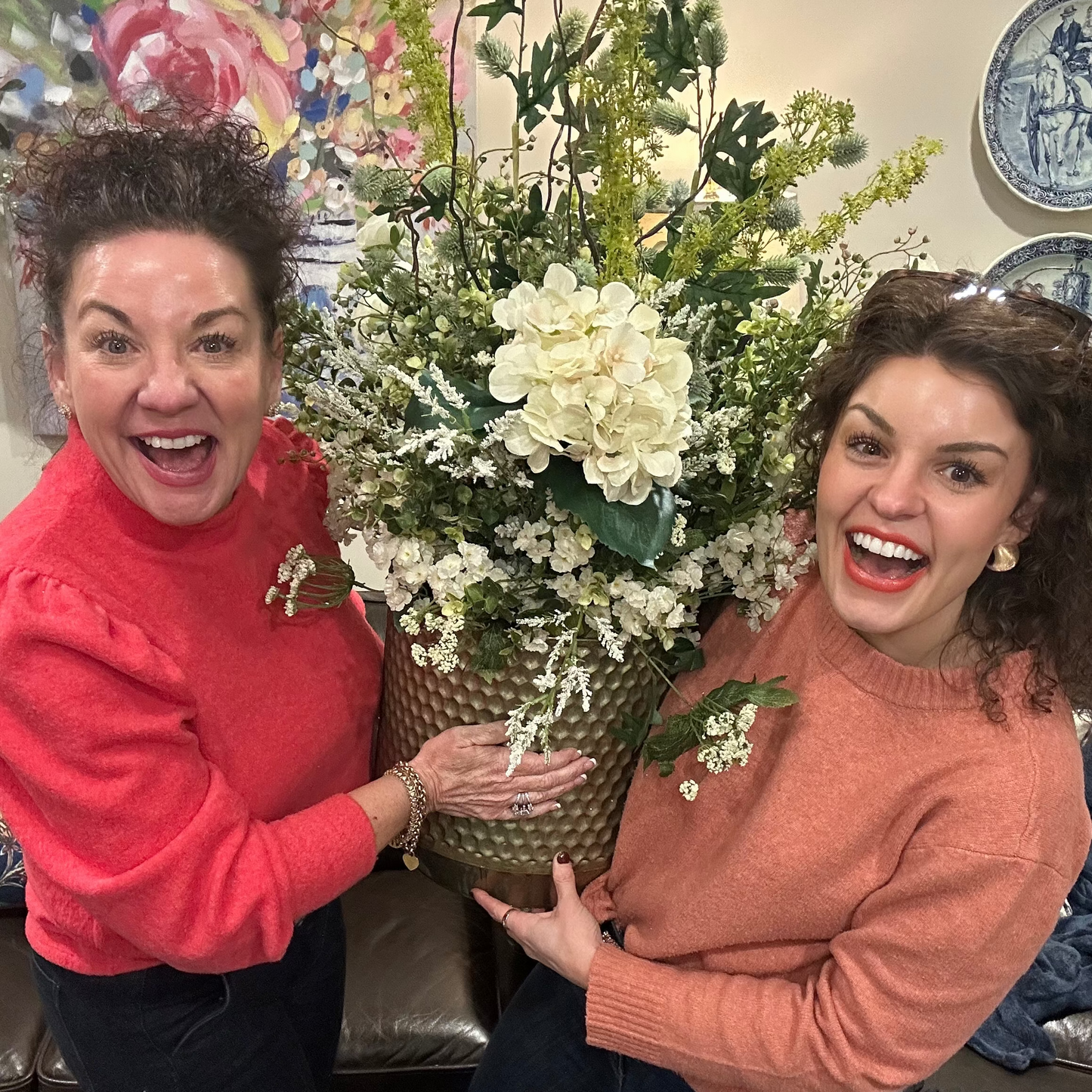 Two women holding a large potted plant in a room with decorative elements.