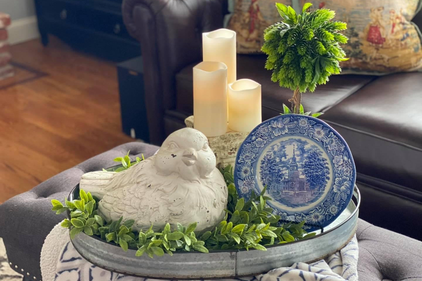 Decorative tray with large bird figurine, candlesticks, faux greenery, and a blue and white vintage/antique plate on an ottoman in the living room.